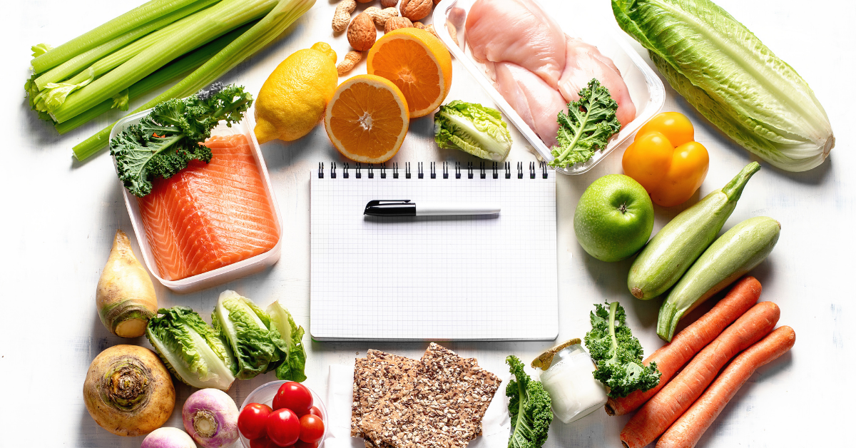notebook and pen on a table surrounded by raw produce, fish, and chicken