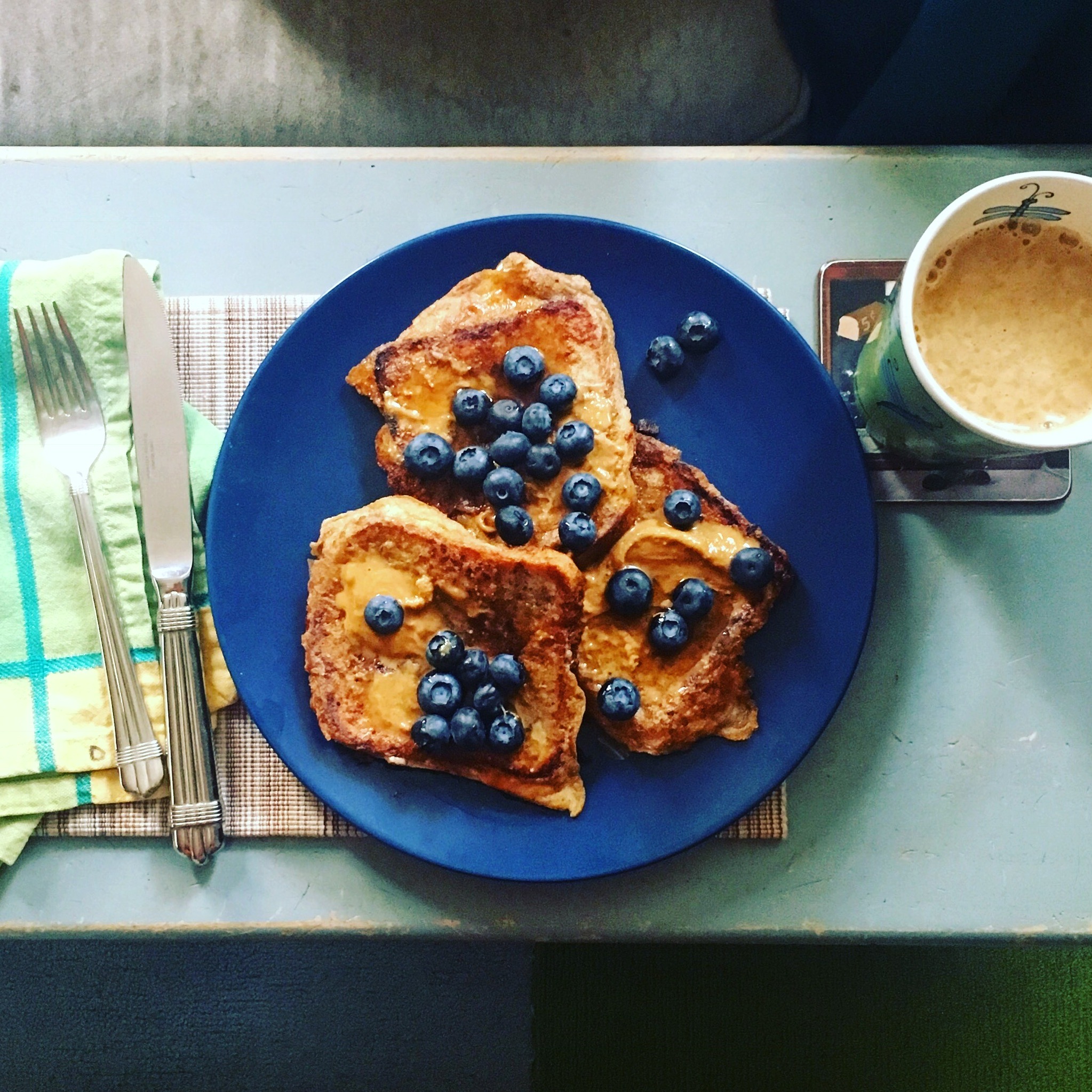 French totast topped with syrup and blueberries on a deep blue plate against a light blue table, with a green napkin to the left and a cup of coffee to the right