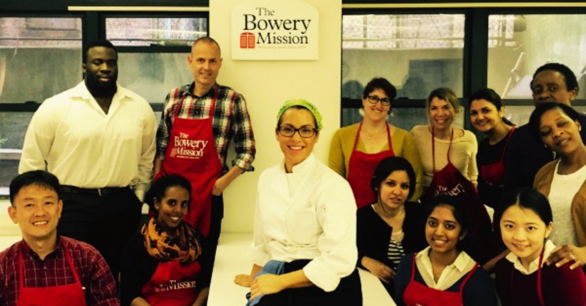 chef sitting on a table facing camera and smiling with volunteers in red aprons flanking her