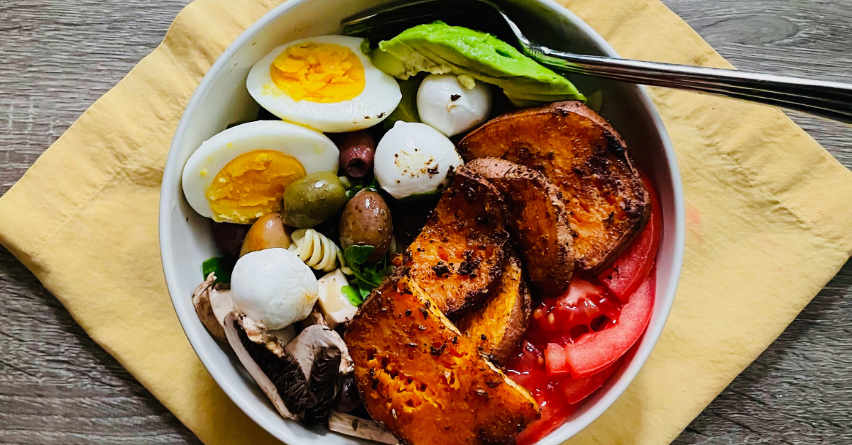 Bowl featuring hard-boiled eggs, cheese balls, roasted sweet potatoes, tomatoes, and avocado