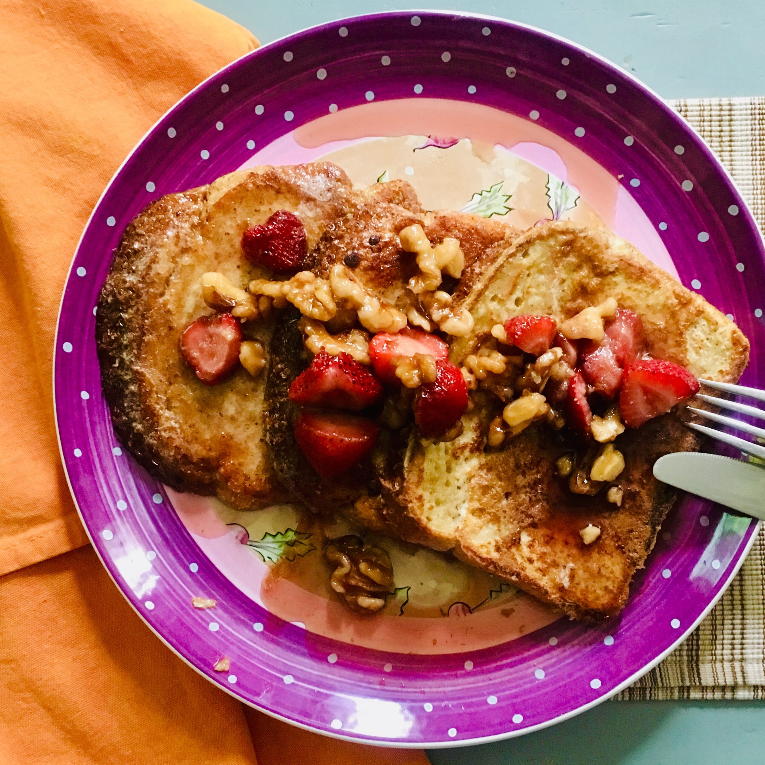 Three slices of French toast with berries and nuts atop a purple-rimmed plate and orange napkin