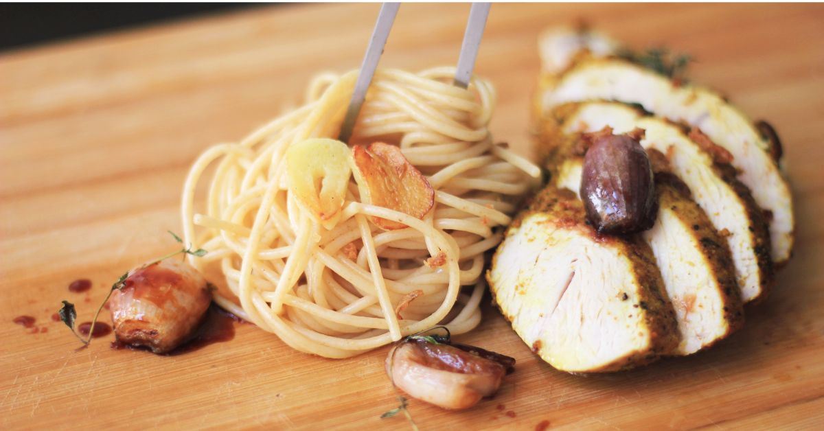Roasted cloves of garlic, a forkful of spaghetti, and sliced grilled chicken sit atop a wooden cutting board. This represents foods that would be helpful in managing inflammatory bowel disease.