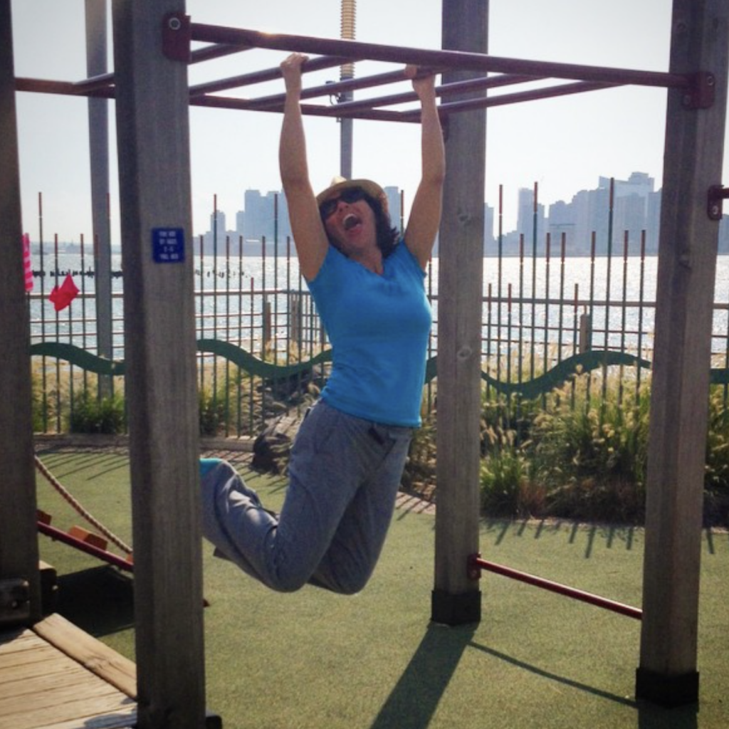 Dina in a hat, blue top, and grey pants, swinging on a jungle gym in a playground.