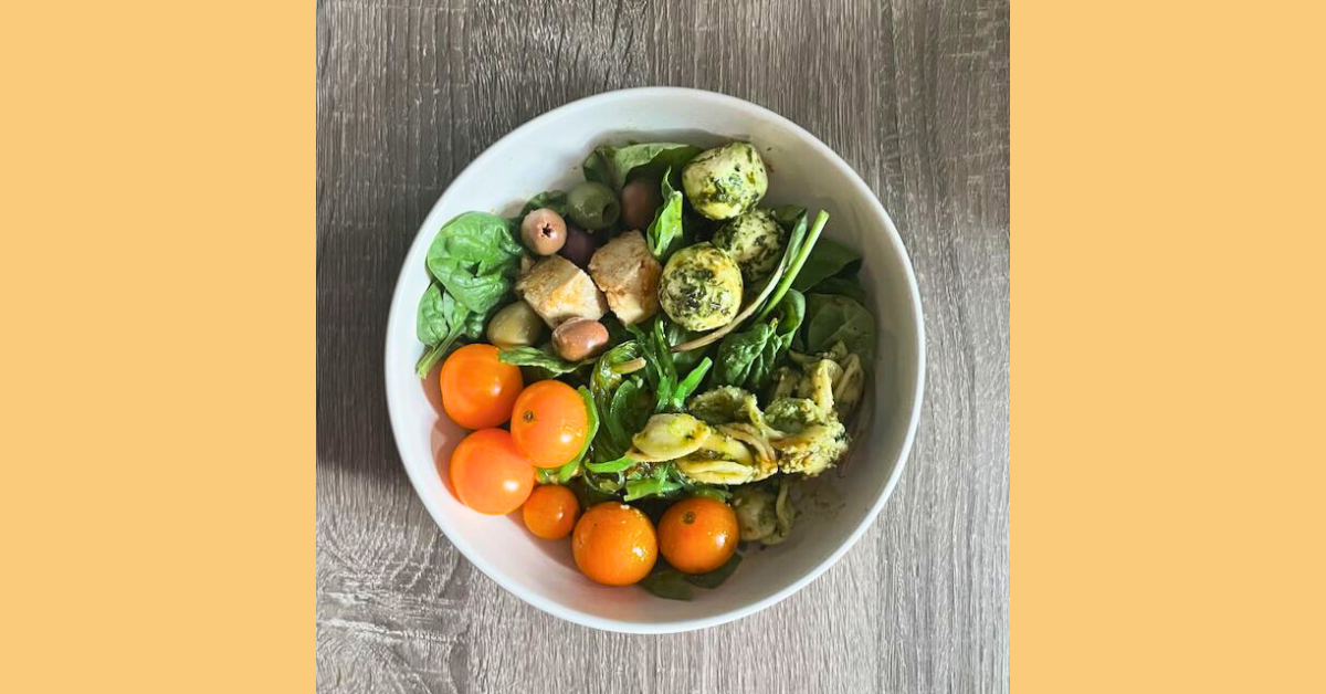 A white bowl full of leafy greens, grilled tofu, and mini orange tomatoes on top of a wood table.