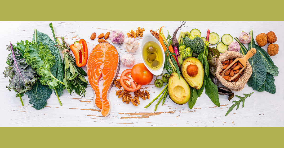 A lineup along a table of heart-healthy foods: leafy greens, salmon fillet, nuts & avocado