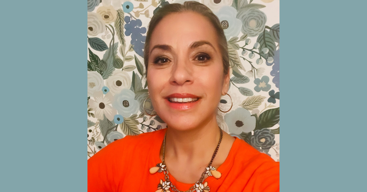 Dina appears in a bright orange top against a floral-patterned wallpaper