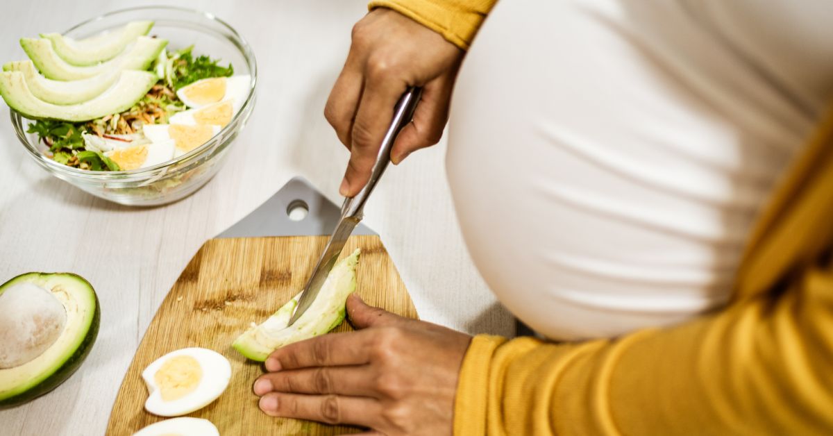 A pregnant person's belly and hands are in view. They are wearing a white top and yellow sleeves while slicing avocado on a cutting board.