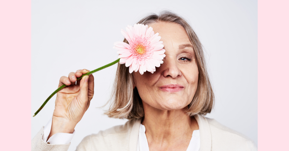 A white woman with a short blond bob haircut is smiling and holding a pink gerbera daisy in front of her right eye