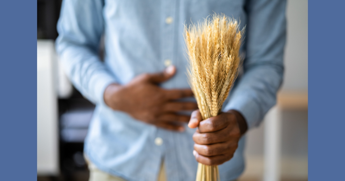 torso of black person in a blue denim shirt clutching their tummy with right hand and extending sheaf of wheat with left hand